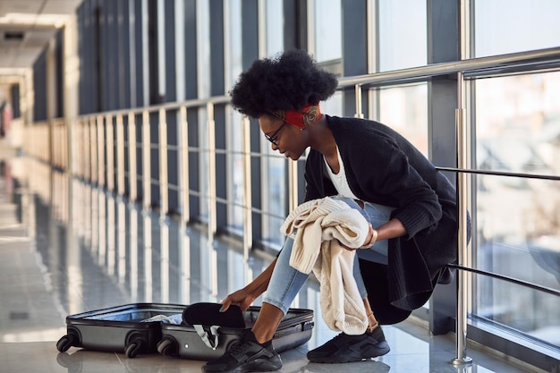 Una joven pasajera afroamericana con ropa informal está en el aeropuerto con ropa para empacar equipaje.