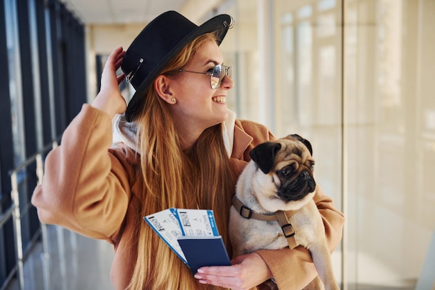 Joven pasajera con ropa de abrigo con boletos y un lindo perro en las manos en el aeropuerto.