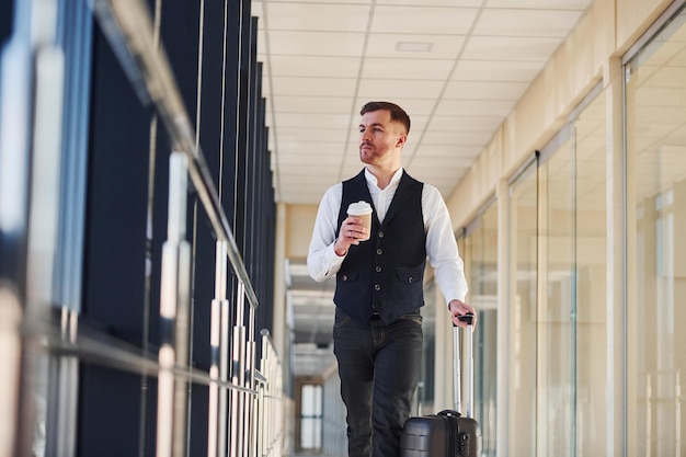 Un joven pasajero con elegante ropa formal está en el vestíbulo del aeropuerto caminando con equipaje y una taza de bebida.