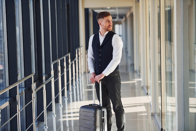 Un joven pasajero con elegante ropa formal está en el vestíbulo del aeropuerto con equipaje.