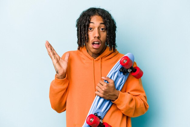 Joven patinador afroamericano aislado de fondo azul sorprendido y conmocionado