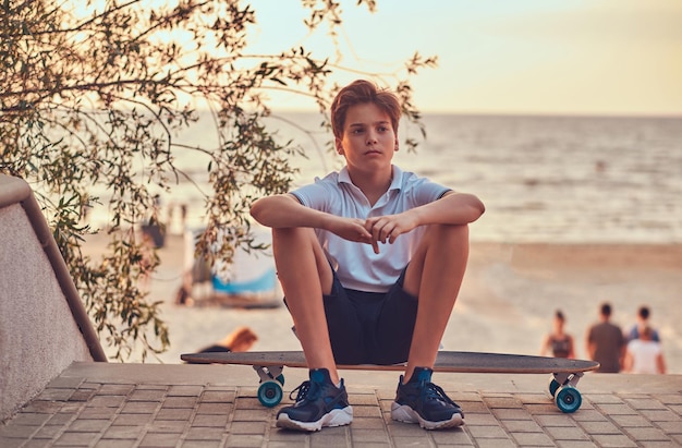 Joven patinador sentado en una patineta contra el fondo de la costa al atardecer.