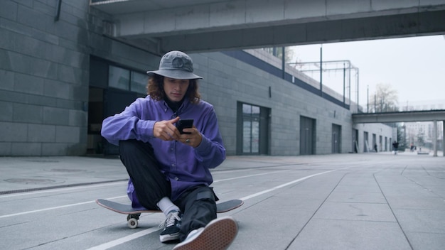 Foto joven patinador sentado en patineta con el teléfono en la mano hipster deportivo con patineta usando un teléfono inteligente al aire libre cool skater mirando a la pantalla del teléfono móvil en el estadio de la ciudad