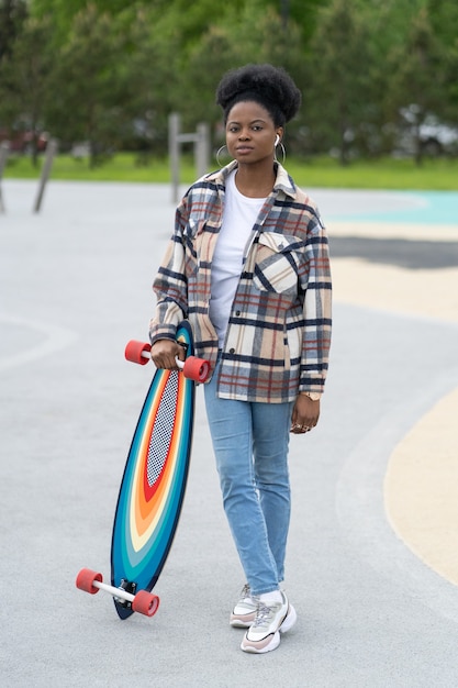 Joven patinadora africana mantenga longboard en skate park mujer urbana en ropa casual en skatepark