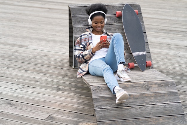 Joven patinadora negra mantenga el texto del teléfono inteligente sentarse en un banco en el espacio urbano con mensajes de longboard