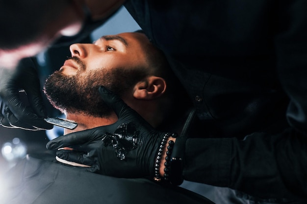 Joven con peinado elegante sentado y afeitándose la barba por un tipo con máscara protectora negra en la barbería