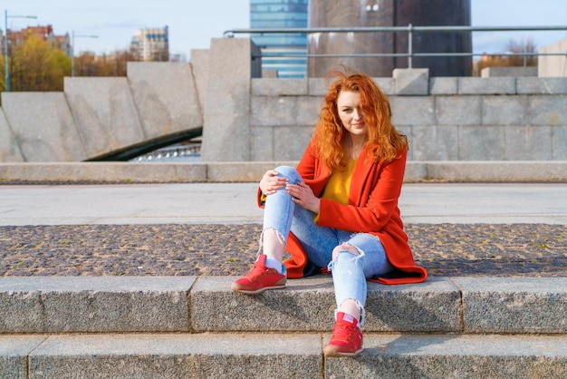 Joven pelirroja con abrigo rojo suéter amarillo y jeans se sienta en las escaleras