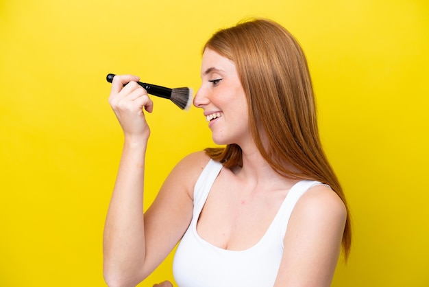 Joven pelirroja aislada de fondo amarillo sosteniendo un pincel de maquillaje y con expresión feliz