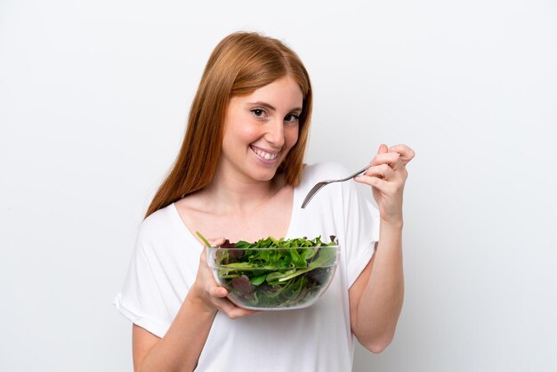 Joven pelirroja aislada de fondo blanco sosteniendo un tazón de ensalada con expresión feliz