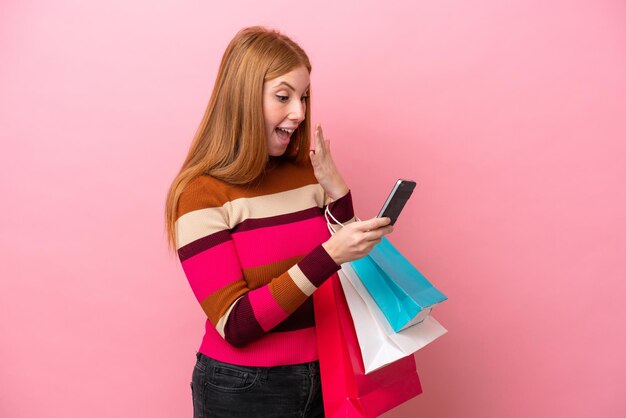 Joven pelirroja aislada de fondo rosa sosteniendo bolsas de compras y escribiendo un mensaje con su teléfono celular a un amigo