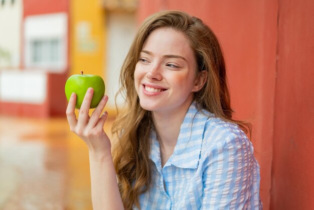 Joven pelirroja al aire libre sosteniendo una manzana con expresión feliz