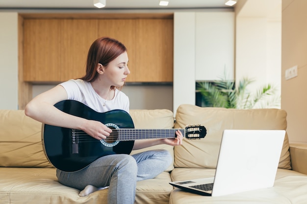 Joven pelirroja aprende a tocar la guitarra con la ayuda de lecciones de video tutorial. mujer mujer en casa en el ocio sentado en el sofá con el portátil estudiando instrumentos musicales en línea. Distancia de pasatiempos