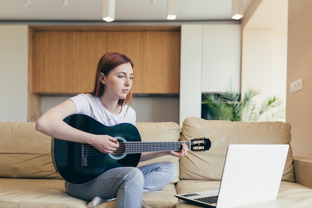 Joven pelirroja aprende a tocar la guitarra con la ayuda de lecciones de video tutorial. mujer mujer en casa en el ocio sentado en el sofá con el portátil estudiando instrumentos musicales en línea. Distancia de pasatiempos