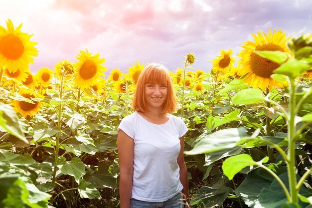 Una joven pelirroja con una camiseta blanca y jeans sonríe y disfruta de un paseo de verano en un campo de girasoles contra un cielo azul en un cálido día de verano