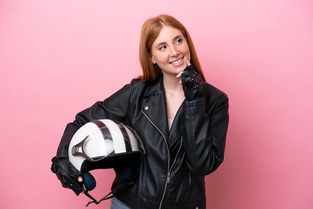 Joven pelirroja con un casco de motocicleta aislado en un fondo rosa pensando en una idea mientras mira hacia arriba