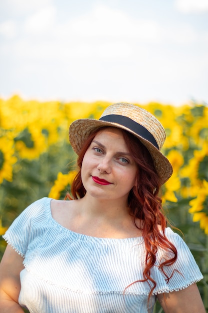 Joven pelirroja chica caucásica en verano vestido de algodón y sombrero de paja en un campo de girasoles amarillos en un día soleado.