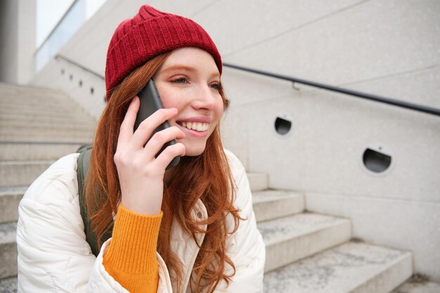 Joven pelirroja con estilo con sombrero rojo se sienta en la calle y habla por teléfono móvil tiene conversaciones telefónicas