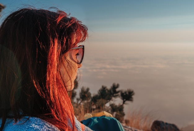 Joven pelirroja con gafas de sol sentada en una roca en lo alto de las montañas por encima de las nubes y