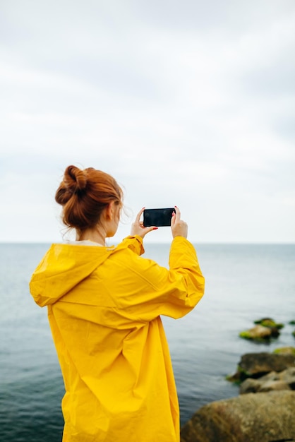 Joven pelirroja con impermeable amarillo posando en la costa del océano tomando fotos de la naturaleza con el teléfono inteligente