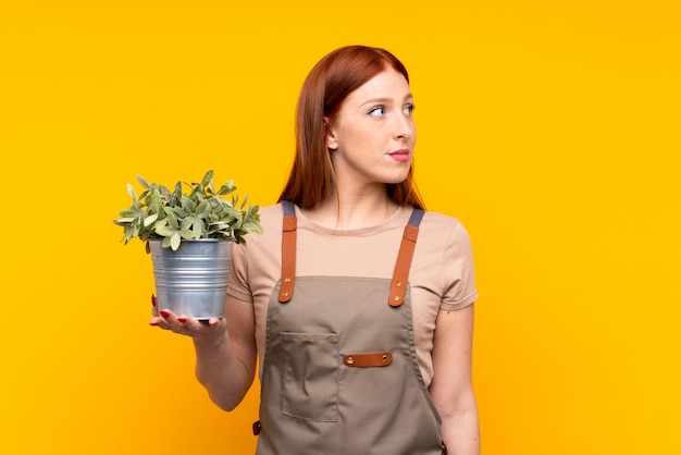 Foto joven pelirroja jardinero mujer sosteniendo una planta sobre pared amarilla aislada mirando hacia el lado