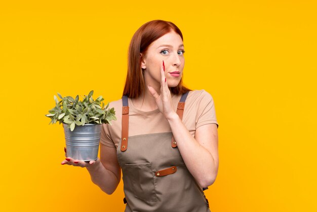 Joven pelirroja jardinero mujer sosteniendo una planta sobre pared amarilla aislada susurrando algo