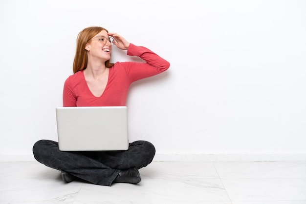 Una joven pelirroja con una laptop sentada en el suelo aislada de fondo blanco se ha dado cuenta de algo y tiene la intención de encontrar la solución