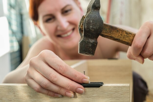 Foto joven pelirroja martillando en un clavo y sonriente mujer recoge muebles quehaceres domésticos bricolaje