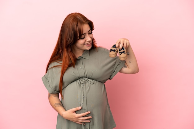 Joven pelirroja mujer caucásica aislada sobre fondo rosa embarazada y sosteniendo botitas de bebé
