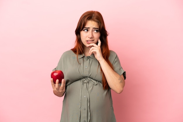 Joven pelirroja mujer caucásica aislada sobre fondo rosa embarazada y sosteniendo una manzana mientras piensa