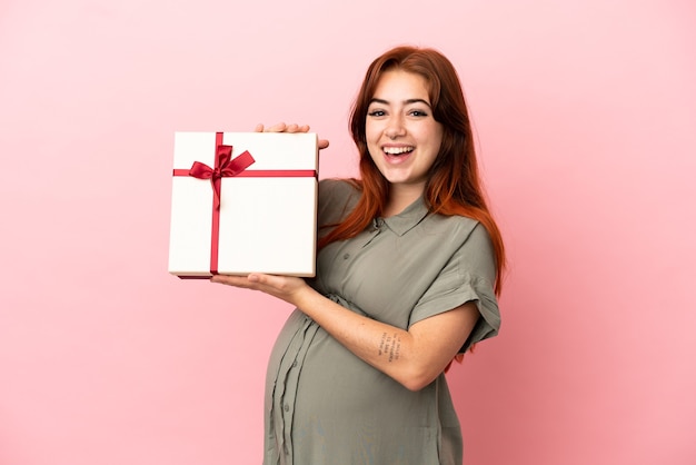 Joven pelirroja mujer caucásica aislada sobre fondo rosa embarazada y sosteniendo un regalo