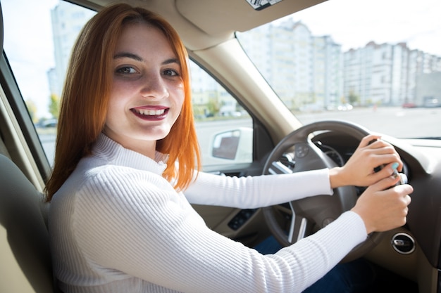 Joven pelirroja mujer conductora detrás de una rueda conduciendo un coche sonriendo felizmente.