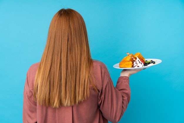 Joven pelirroja mujer sosteniendo waffles sobre pared aislada en posición posterior