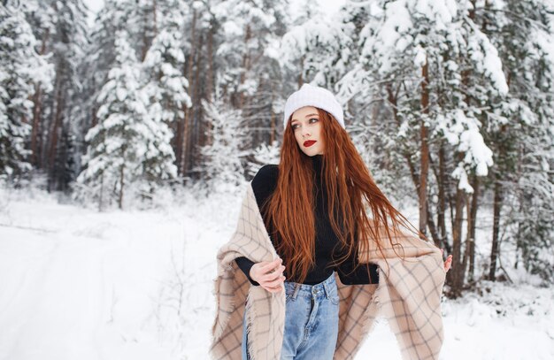 Joven pelirroja con pelo largo rojo sobre un fondo de invierno. Mujer pelirroja con un sombrero blanco en el fondo de la naturaleza de invierno.