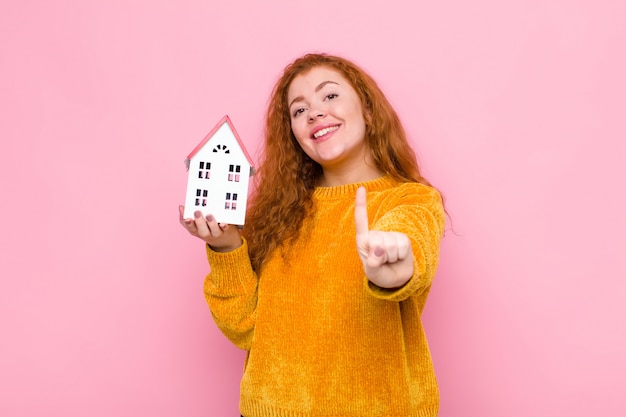 Foto joven pelirroja que sonríe con orgullo y confianza haciendo que la número uno pose triunfalmente, sintiéndose como una líder con una modelo de casa