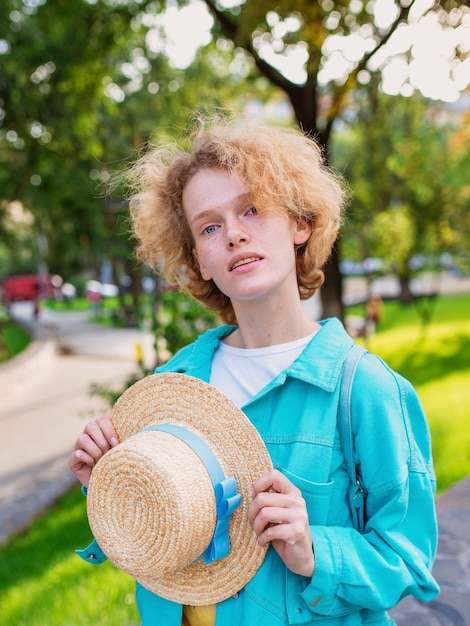 Joven pelirroja rizada alegre mujer en chaqueta azul sosteniendo sombrero de paja en sus manos y feliz