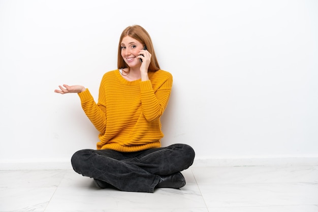 Joven pelirroja sentada en el suelo aislada de fondo blanco manteniendo una conversación con alguien por teléfono móvil