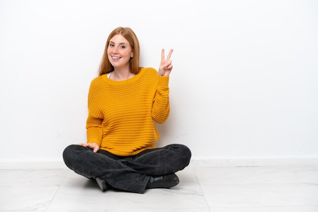 Foto joven pelirroja sentada en el suelo aislada de fondo blanco sonriendo y mostrando el signo de la victoria