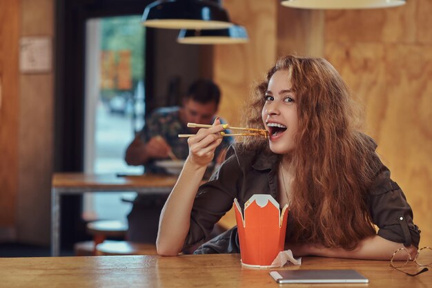 Una joven pelirroja sonriente con ropa informal comiendo fideos picantes en un restaurante asiático.