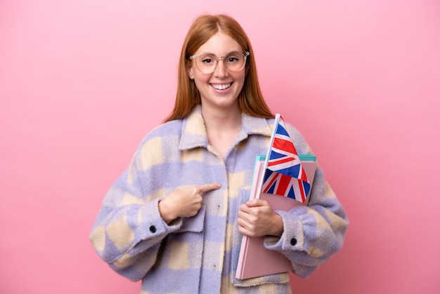 Joven pelirroja sosteniendo una bandera del Reino Unido aislada de fondo rosa con expresión facial sorpresa