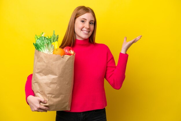 Joven pelirroja sosteniendo una bolsa de compras aislada en un fondo amarillo extendiendo las manos a un lado para invitar a venir