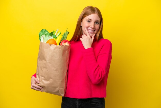 Joven pelirroja sosteniendo una bolsa de compras aislada de fondo amarillo feliz y sonriente