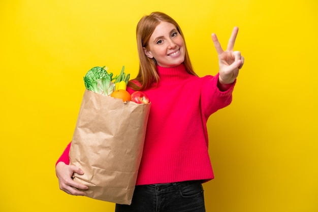 Joven pelirroja sosteniendo una bolsa de compras aislada de fondo amarillo sonriendo y mostrando el signo de la victoria