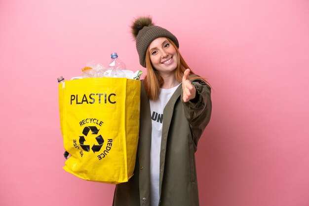 Joven pelirroja sosteniendo una bolsa llena de botellas de plástico para reciclar aisladas en un fondo rosado dándose la mano para cerrar un buen trato