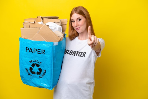 Joven pelirroja sosteniendo una bolsa de reciclaje llena de papel para reciclar aislada de fondo amarillo mostrando y levantando un dedo