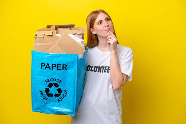 Joven pelirroja sosteniendo una bolsa de reciclaje llena de papel para reciclar aislada en un fondo amarillo que tiene dudas mientras mira hacia arriba