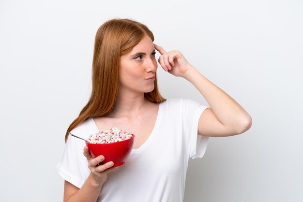 Joven pelirroja sosteniendo un tazón de cereales aislado de fondo blanco que tiene dudas y con una expresión facial confusa