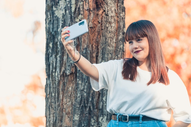 Joven pelirroja tomando un selfie, transmisión de video en línea en el parque durante una temporada otoñal. Camiseta blanca blue jeans traje moderno de moda. Red social, creador de contenido con espacio de copia.