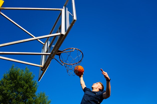 Joven pelirrojo con una camiseta azul oscuro lanza una pelota en movimiento en un aro de baloncesto contra un cielo azul al aire libre