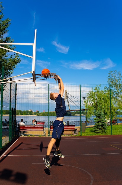 Joven pelirrojo con una camiseta azul oscuro lanza una pelota en movimiento en un aro de baloncesto contra un cielo azul al aire libre