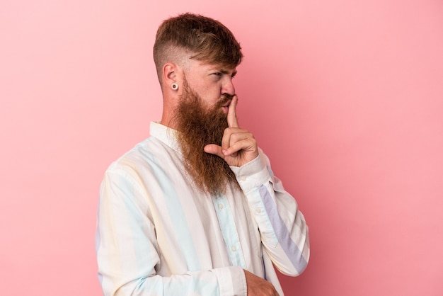 Joven pelirrojo caucásico con barba larga aislado sobre fondo rosa guardando un secreto o pidiendo silencio.